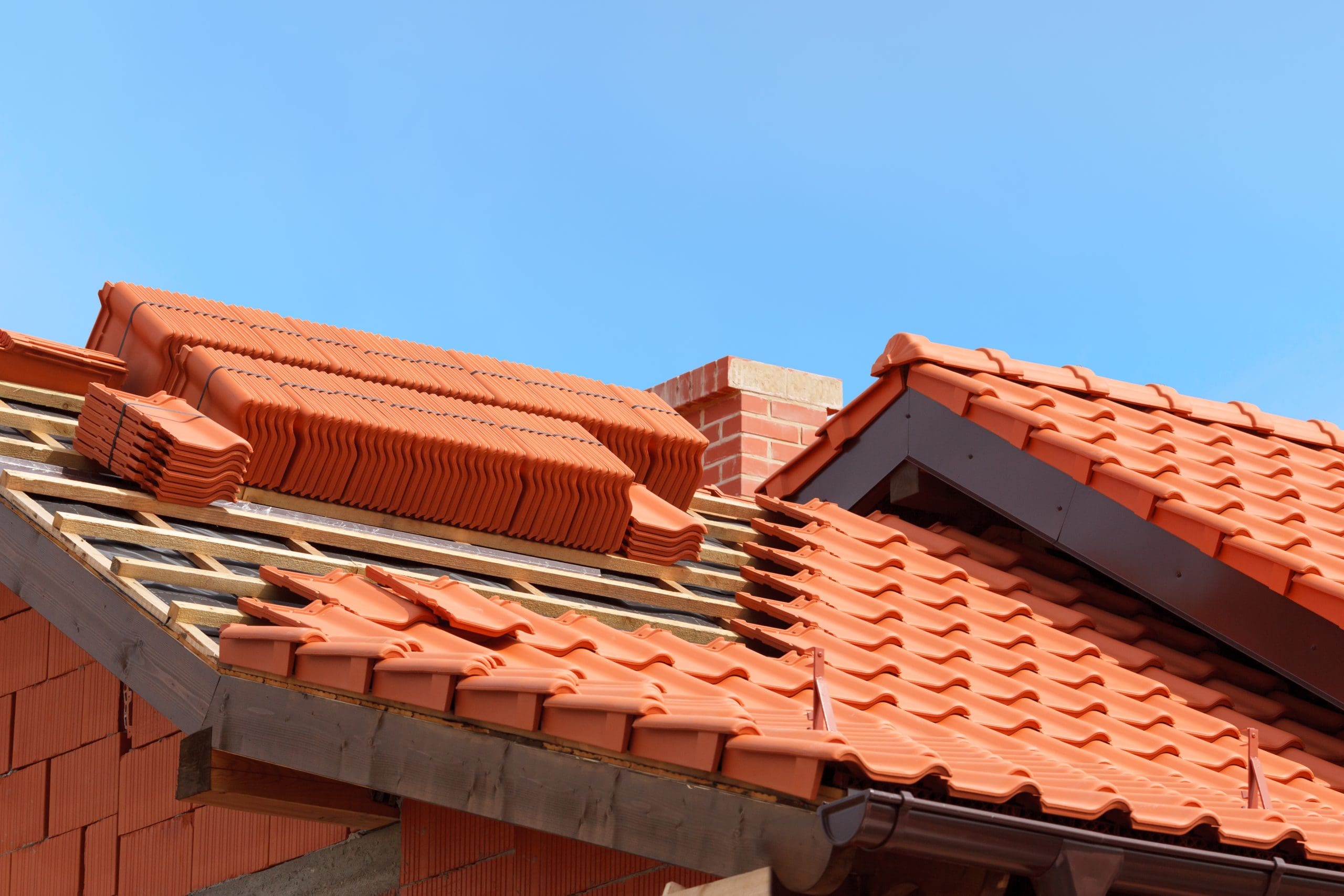 A newly constructed house roof is shown with stacked red terracotta tiles. Some tiles are neatly arranged on the roof, while a bundle remains uninstalled. A brick chimney is visible under a clear blue sky.