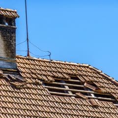 A damaged tile roof with missing and dislodged tiles, exposing wooden beams underneath, set against a clear blue sky. A chimney with aged brickwork is visible, and a metal antenna is mounted on the roof.
