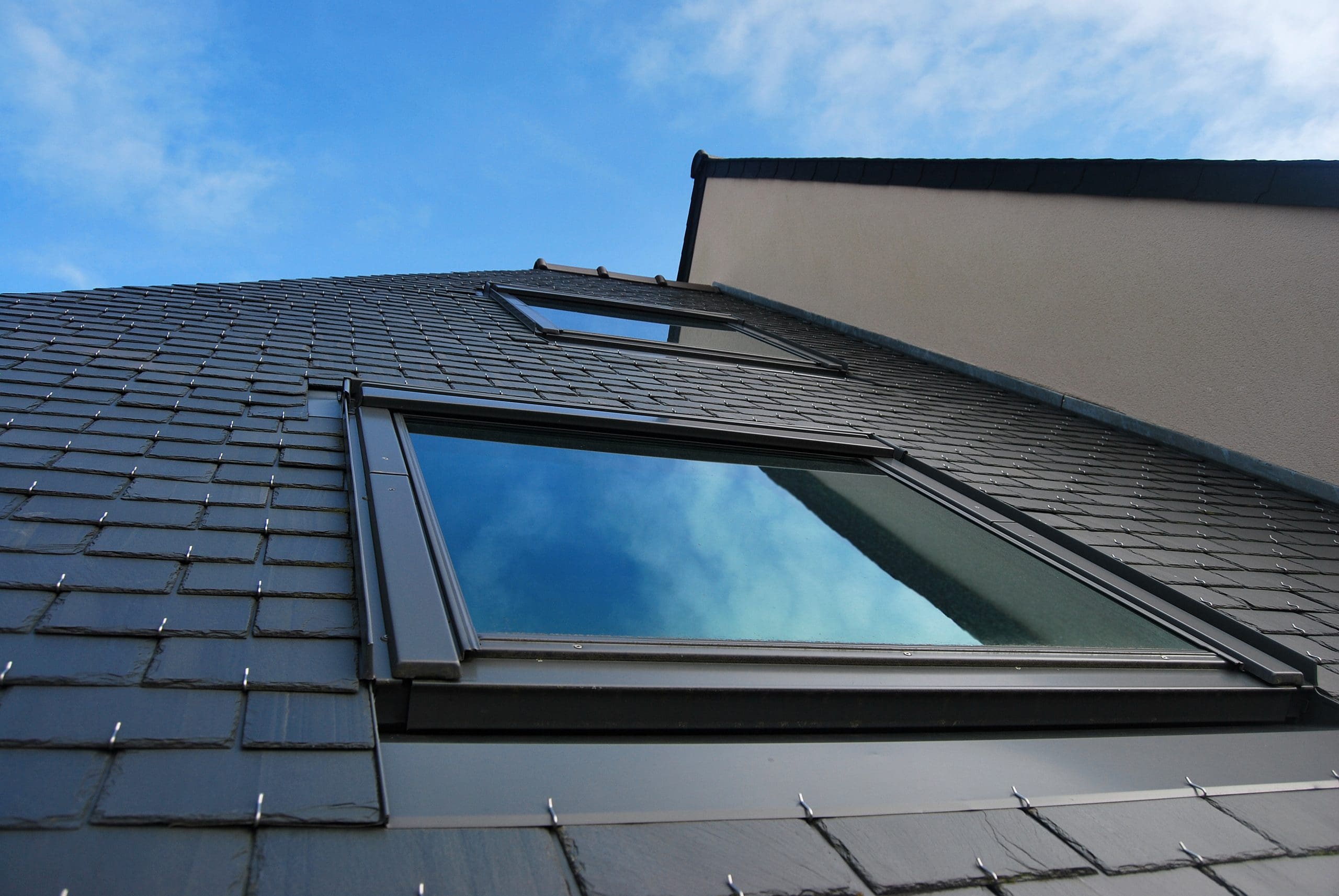 Close-up view of a modern house with dark slate tiles and large reflective windows, set against a bright blue sky. The unique angle emphasizes the sleek, contemporary design and clean lines of the structure.