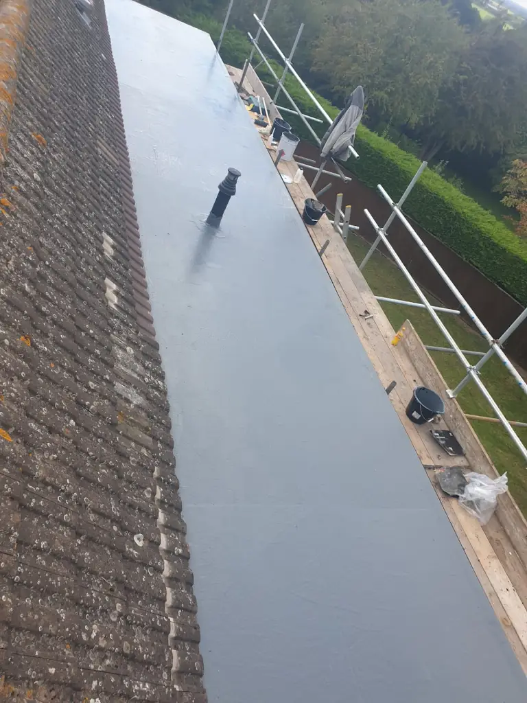 View of a flat roof under construction, with a newly applied gray surface. Scaffolding surrounds the area, and various construction tools and materials are visible. A sloped tiled roof is adjacent, with trees in the background.