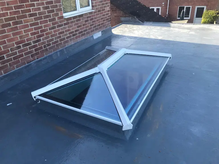 A pyramid-shaped skylight on a flat rooftop, surrounded by brick walls and additional flat-roofed structures. The skylight has a metal frame and clear glass panels that allow sunlight to illuminate the interior space below.
