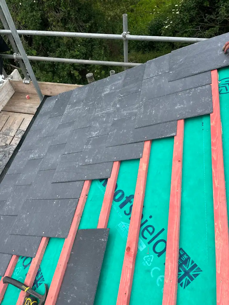 Partially constructed roof with overlapping dark slate tiles on a green underlayment. Red wooden battens are visible beneath the tiles. A scaffolding and greenery are in the background.