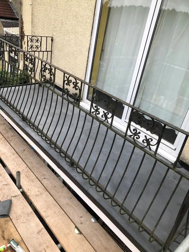 A balcony with decorative wrought iron railings is seen from above. The balcony floor appears new, contrasting with the older, ornate railings. A glass door with white curtains is in the background, and wooden planks are in the foreground.