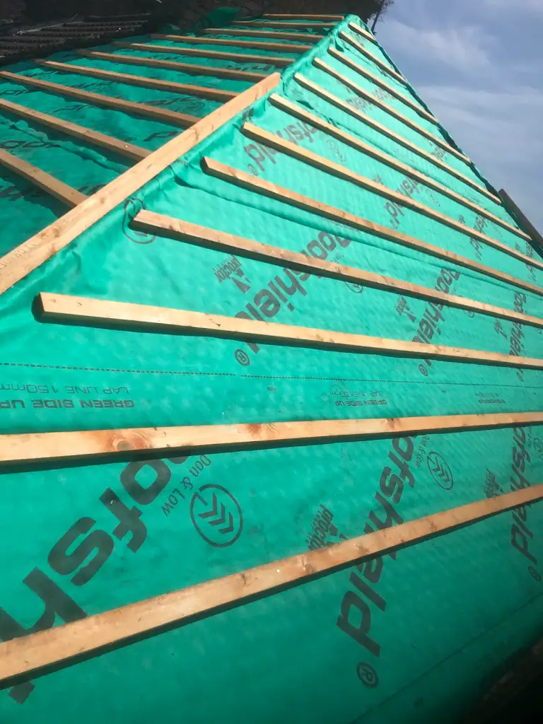 A roof under construction covered with a green waterproof membrane. Wooden battens are placed diagonally across the surface, preparing for the next layer of roofing material against a partly cloudy sky.